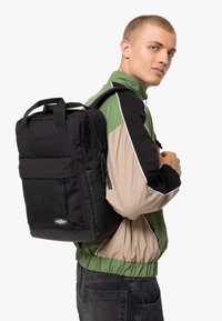 Svart ryggsäck med tyg- och syntetmaterial; har en framficka, dubbla dragkedjor och korta handtag. Bärs på ryggen.