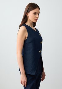 Gilet in lino blu navy con scollatura a forma curva, caratterizzato da tre bottoni oro a forma di conchiglia e una vestibilità rilassata. Tessuto morbido senza decorazioni.
