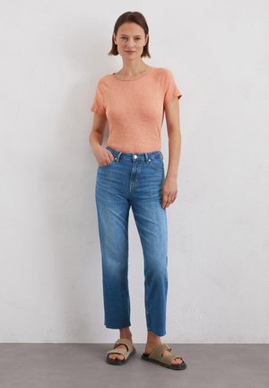 Light orange short-sleeve top with a relaxed fit, paired with straight-leg blue jeans and simple brown sandals on a neutral background.