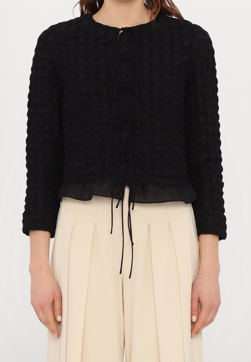 Femme portant un haut noir texturé à manches longues avec des liens sur le devant et un pantalon plissé beige taille haute sur fond blanc.