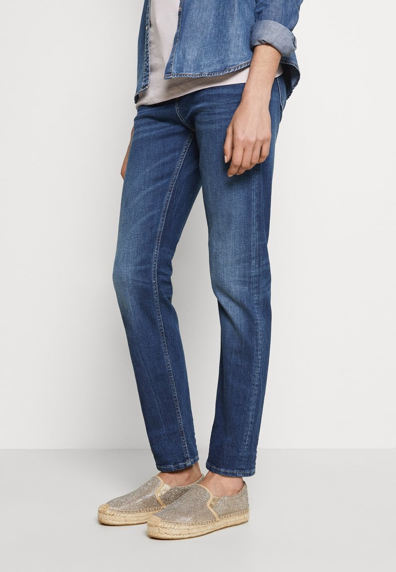 Personne portant un jean slim bleu, une chemise en denim aux manches retroussées, et des espadrilles à enfiler pailletées sur un fond clair.