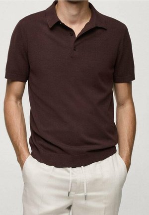 Mann trägt ein dunkelbraunes Poloshirt mit kurzen Ärmeln, das in hellbeige Kordelzughosen gesteckt ist, Hände in den Taschen.