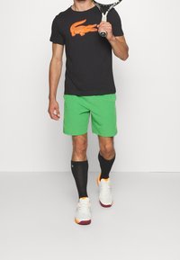 Black t-shirt with orange crocodile logo, green shorts, black knee-length socks, and white-orange sneakers with maroon soles.