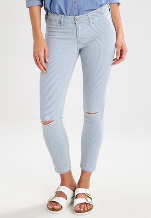 Woman wearing light blue and white striped ripped skinny jeans and white buckle sandals, standing against a plain background.