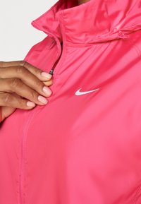 Lyserød nylon zip-jakke med høj krave, med et lille hvidt Nike-logo på brystet og synlige syninger nær lynlåsen.