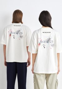 Kremfarvede bomulds t-shirts med tryk af "MILANO" og "MCMLXXVIII" i sort, samt flerfarvede stænk mønstre på ryggen.