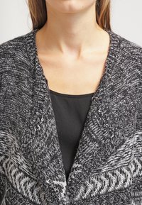 Stickad cardigan i svart och grått, med ett strukturerat mönster och bred krage. Buren över en svart topp, med en lös passform.