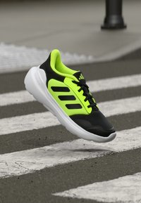 Schwarzer und neongrüner Sportschuh mit einem Obermaterial aus Mesh, drei schwarzen Streifen, gepolsterter Kragen und einer weißen Sohle, vor einem gestreiften Zebrastreifen.