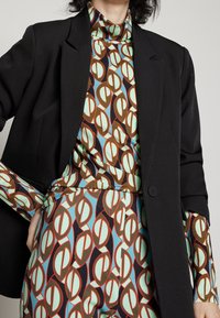 Zwarte blazer over een kleurrijke gebreide trui met een patroon en een broek met verweven vormen in teal, bruin en rood, met lange mouwen.