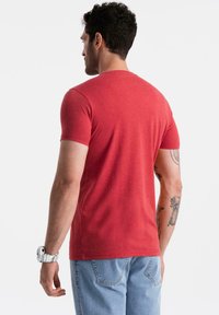 Uomo con capelli corti e ricci, che indossa una maglietta rossa, jeans azzurri chiari, un orologio bianco al polso e tatuaggi visibili sulle braccia, di spalle rispetto alla fotocamera.