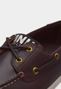 Zbliżenie na ciemnobrązowy skórzany but żeglarski z złotymi przelotkami, skórzanymi sznurowadłami oraz białym napisem na czarnej metce na języku.