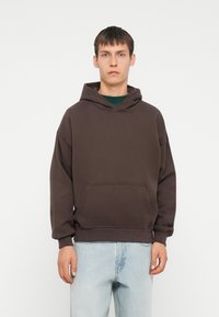 Brun hættetrøje med kængurulomme, snørret hætte og ribbede manchetter. Båret over en grøn skjorte, parret med lyseblå jeans.