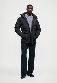 Μαύρο φουσκωτό μπουφάν με κουκούλα, με φερμουάρ και κουμπιά, δύο μπροστινές τσέπες, φορεμένο πάνω από ελαφρύ γκρι πουλόβερ, συνδυασμένο με σκούρα τζιν παντελόνι.