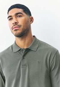 Tricou polo verde măsliniu, realizat dintr-un amestec de bumbac, având o plachetă cu două nasturi, mâneci scurte și un mic logo brodat pe piept.