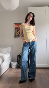 Mlada ženska stoji v spalnici, oblečena v rumeno majico brez rokavov, široke modre kavbojke in črne čevlje, z rokami v žepih.