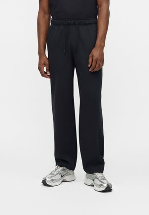 Homme portant un pantalon noir large à taille cordon et des baskets Nike argentées, debout devant un fond blanc uni.