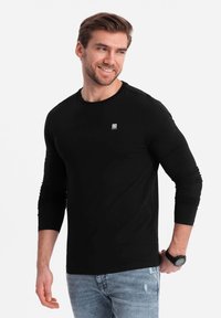 Schwarzes Langarmshirt mit Rundhalsausschnitt und kleinem Logo auf der Brust. Getragen mit hellblauen, verwaschenen Jeans. Glatte Textur.
