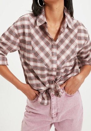Kvinde iført en lyserød og brun ternet skjorte med knapper, bundet om livet, med lyserøde højtaljede jeans og store store øreringe.