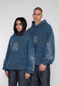 Sweatshirts bleus à coupe ample, avec un logo central et un texte graphique sur les manches. Fabriqués en tissu texturé avec un design décontracté.