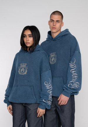 Sudaderas azules de corte holgado, con un logo central y texto gráfico en las mangas. Fabricadas con una tela texturizada y un diseño relajado.