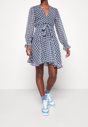 Persona que lleva un vestido de cuadros azul y blanco con mangas largas transparentes, cintura ceñida y zapatillas blancas con detalles azules y calcetines a juego.