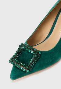 Scarpa flat in suede turchese con dettaglio di grande fibbia quadrata in gemma smeraldo sulla parte superiore anteriore.