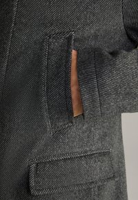 Grau strukturierter Mantel mit Reverskragen, einer seitlichen Tasche mit Klappe und sichtbaren Nähten. Das Material scheint weich und dick zu sein.