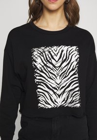 Sweatshirt preto cropped com um gráfico de faixa de zebra branca na frente. Apresenta um decote arredondado e uma bainha desfiada para um visual casual.