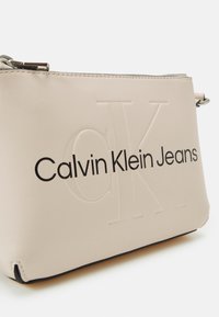 Borsa in faux leather beige con logo "CK" in rilievo, testo nero "Calvin Klein Jeans," chiusura con zip e trim nero alla base.