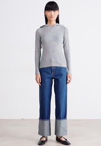 Kvinne som står iført en langermet stripete hettegenser, brede jeans med oppbrett i blått og svarte ballettsko på en ensfarget bakgrunn.