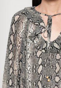 Gjennomsiktig bluse med slangerutemønster, med volangkrage, knyting og metalliske detaljer. Overveiende fargene svart, beige og hvit.
