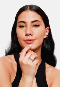 Anello d'argento con una fascia testurizzata indossato sulla mano destra; modella con lunghi capelli scuri che tocca delicatamente le labbra, indossando un top senza maniche nero.