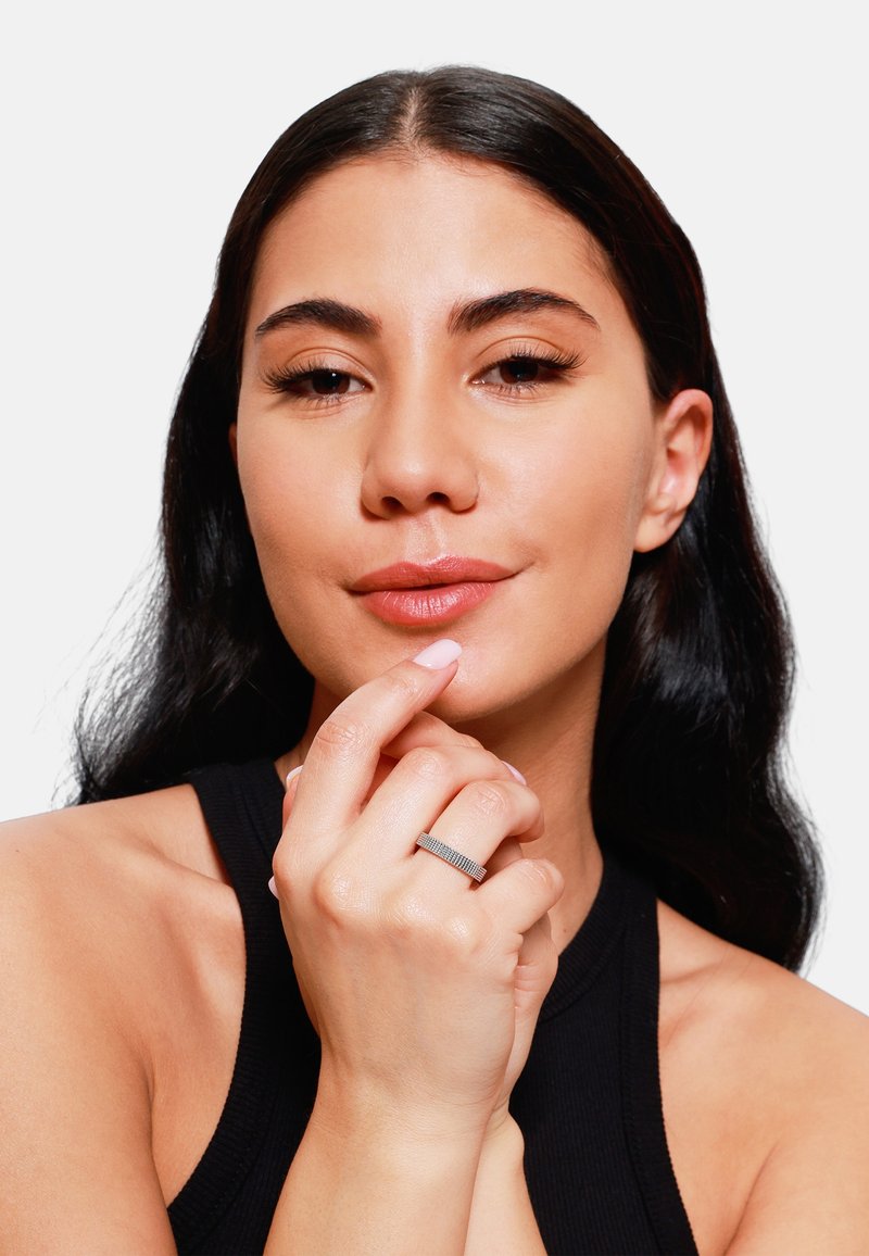 Anello d'argento con una fascia testurizzata indossato sulla mano destra; modella con lunghi capelli scuri che tocca delicatamente le labbra, indossando un top senza maniche nero.