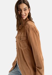 Bruin corduroy overhemd met horizontale ribpatronen, een knopenfront en twee borstzakken. De stof lijkt zacht en heeft een relaxte pasvorm.
