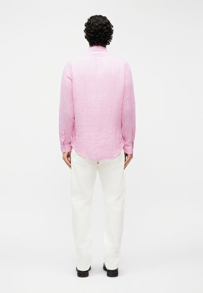 Άνδρας με σγουρά σκούρα μαλλιά, που φοράει μακρυμάνικο ροζ πουκάμισο και λευκό παντελόνι, στέκεται με την πλάτη προς το μέρος του θεατή, πάνω σε απλό φωτεινό φόντο.