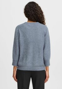 Pull en maille bleue avec une texture côtelée, col rond et manches trois-quarts, associé à un pantalon noir.