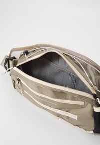 Borsa in tessuto beige con più compartimenti con cerniera, dotata di fodera in rete, tracolla regolabile e dettagli con logo. L'interno presenta tasche.