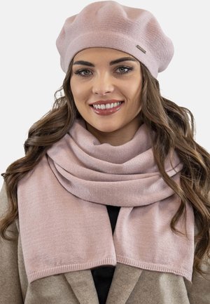 Béret en tricot rose clair associé à une écharpe assortie, présentant une texture lisse et de subtils accents côtelés, porté sur un manteau beige.