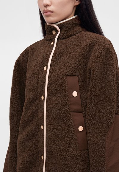 Γυναίκα που φοράει καφέ υφασμένη fleece ζακέτα με μπεζ κουμπιά τύπου snap και ψηλό γιακά, εμφανιζόμενη από το στήθος μέχρι το πιγούνι σε μονόχρωμο φόντο.