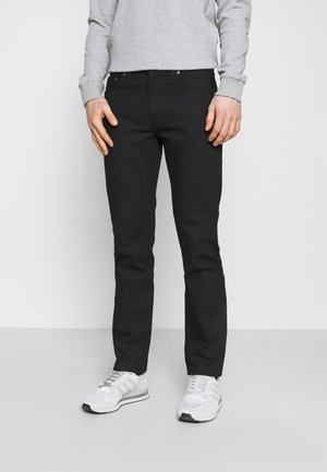 Mann trägt schwarze Slim-Fit-Hose, hellgraues Langarmshirt und weiße Sneaker, steht vor einem schlichten Hintergrund.