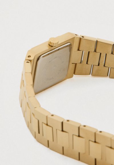Pulseira de relógio em metal dourado com elos rectangulares e caixa do relógio quadrada fechada vista por trás sobre um fundo branco.