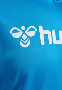 Blå skjorta i tyg med en vit logotyp som har bielement och ordet "hu." Materialet verkar smidigt med en lätt textur.