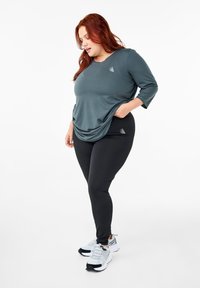 Dark teal long-sleeve athletic top with a draped design and logo, paired with black leggings featuring a logo. White athletic shoes complete the look.