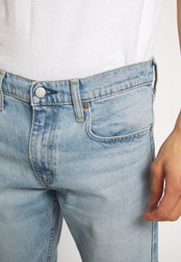 Gros plan d'une personne portant un jean en denim bleu clair et une chemise blanche, avec la main droite détendue sur le côté.