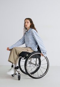 Blå och vit randig långärmad skjorta, beige vida byxor, sittande i en metallisk rullstol med sporthjul på en enfärgad bakgrund.