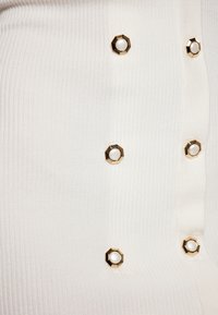 Tissu blanc à côtes avec une rangée de boutons hexagonaux dorés ornés de centres en perles blanches sur le côté droit.