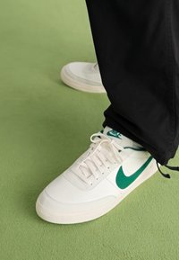 Valged nahast tossud rohelise Nike Swooshiga, tekstureeritud pind, ümmargune varvas ja lame kummitald, koos mustade pükstega.