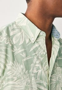 Camicia verde chiaro con motivo a foglie, tessuto lavorato e bottoni bianchi. Colletto a contrasto con dettagli geometrici blu.