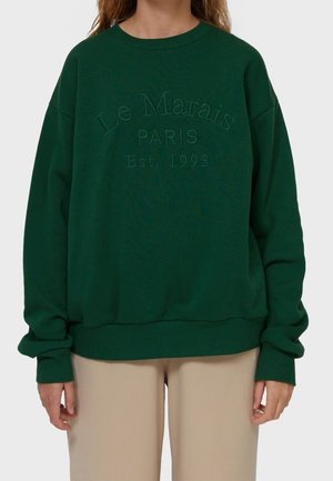 Osoba nosząca ciemnozieloną bluzę z wyembosowanym napisem ""Le Marais PARIS Est. 1993"" na przodzie, w połączeniu z jasnobeżowymi spodniami.