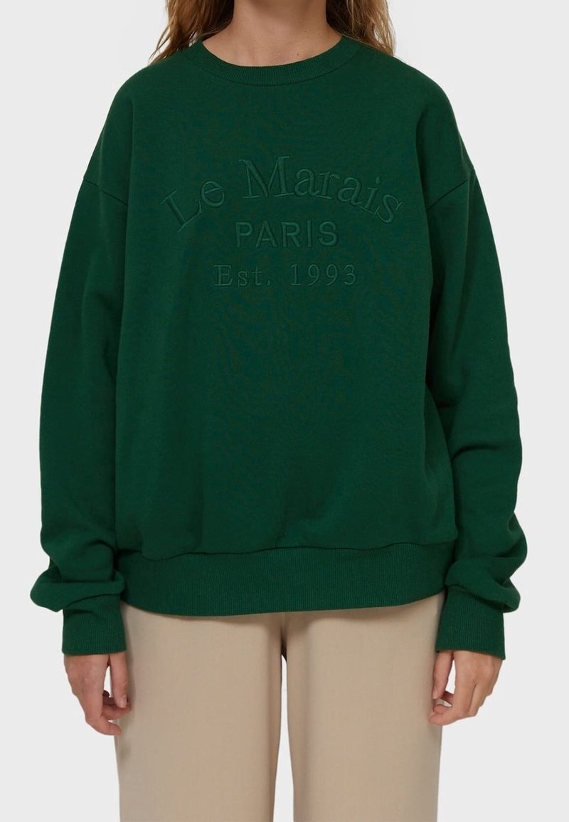 Personne portant un sweat-shirt vert foncé avec "Le Marais PARIS Est. 1993" embossé sur le devant, associé à un pantalon beige clair.